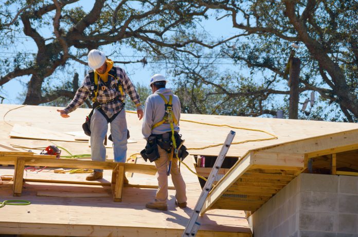 Two construction workers are on top of a tall building wearing safety harnesses that are attached to lifelines to protect them from injury if they fall off the building.