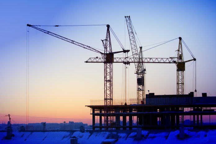 Construction cranes are building a house against the background of sunset and dark sky