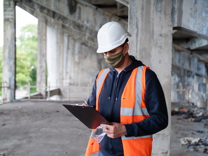 An engineer inspects a construction site in a construction helmet and an antiviral mask during a coronavirus pandemic.