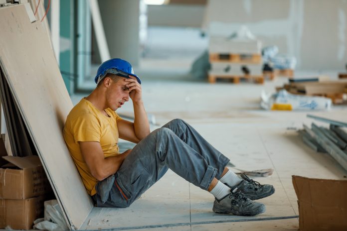 A young Caucasian construction worker is sitting at work in a depressive state, with his hand on his forehead.