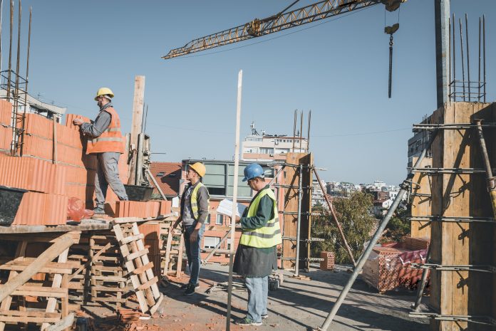Teamwork by Three Builders with Protective Helmets who are Building a Wall of Bricks at the Building Site. Young Building Workers is Working Together at the Roof of the Construction Site Under Control of Their Experienced Team Leader.