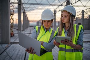 Female construction engineers with helmet are holding laptop and tablet and discussing the work progress on the construction site.