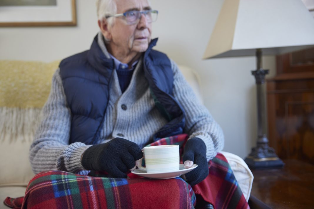 Senior Man Wearing Extra Clothes With Hot Drink Trying To Keep Warm At Home In Energy Crisis