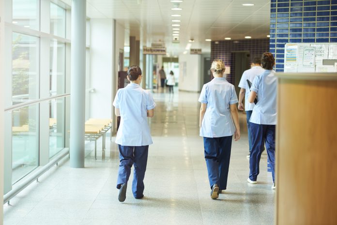 Five major hospitals to be rebuilt as part of £20bn New Hospital Programme a group of four young trainee nurses including male and female nurses , walk away from camera down a hospital corridor . They are wearing uk nurse uniforms of trousers and tunics.
