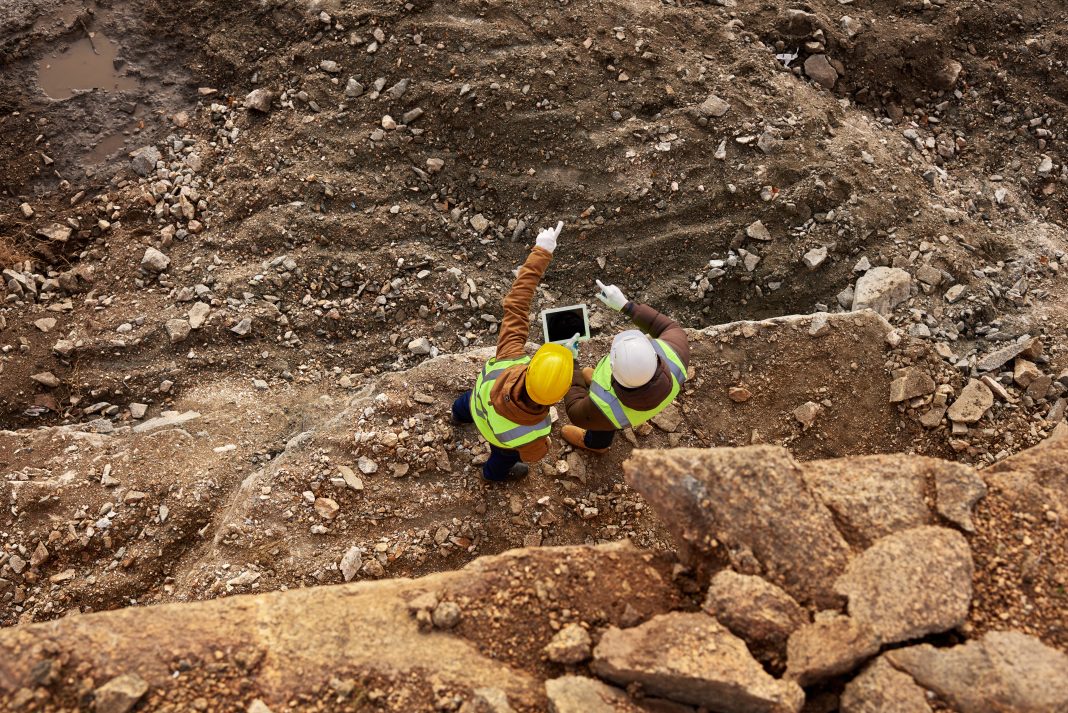 To represent the new Carbon Quantifier from RIB software, a top view shot of two industrial workers wearing reflective jackets standing on mining worksite outdoors using digital tablet, copy space