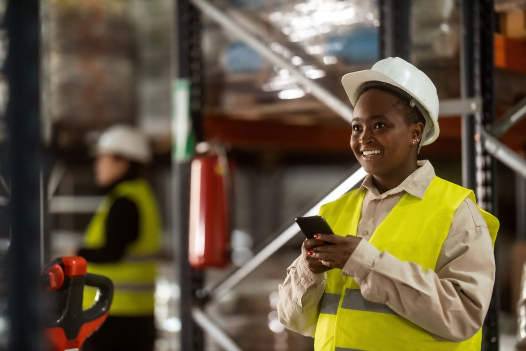 An African-American warehouse worker is using a mobile phone at work with a big smile on her face, representing why a diversity strategy is needed