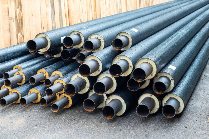 Heap of many new black insulated steel pipes at municipal construction site outdoors. Heating main district pipeline renewal or reconstruction. City development building industrial background, representing BS 5422:2023