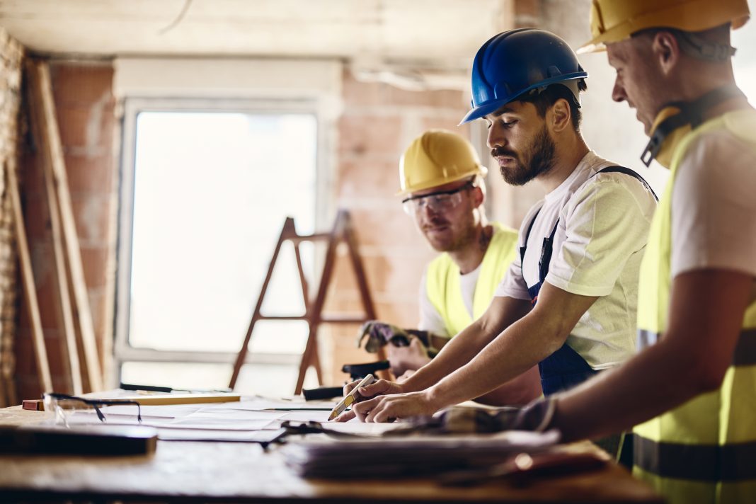 Group of construction workers cooperating while analyzing blueprints at renovation site, representing construction output growth. Focus is on man in the middle.