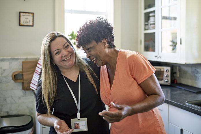 Waist-up view of Black client and healthcare worker standing with arms around each other and laughing during routine house call, representing Supported Housing Design