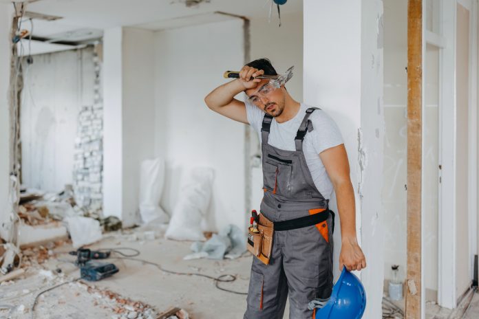 A young Caucasian male construction worker is taking a break to wipe the sweat from his forehead, representing the stress SME housebuilders currently face