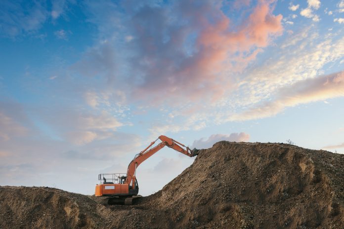 Excavator bulldozer in sandpit with raised bucket over blue cloudscape sky. The latest ONS figures for the month-on-month series show that construction output continues to fall, with a 0.2% decrease in volume terms