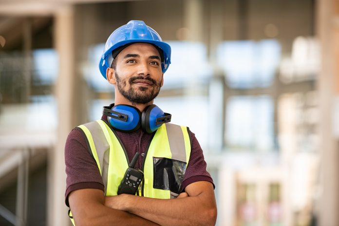 Construction site manager standing with folded arms wearing safety vest and helmet, thinking at construction site. Young architect watching construction site with confidence. Portrait of mixed race manual worker looks at his work with satisfaction.