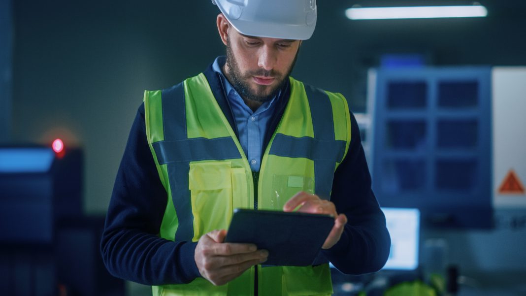 Professional Engineer Wearing Safety Vest and Hardhat Stands in Modern Factory Workshop, Uses Digital Tablet Computer. Handsome Professional Working in Industrial Facility with High Tech Machinery Professional Engineer Wearing Safety Vest and Hardhat Stands in Modern Factory Workshop, Uses Digital Tablet Computer. Handsome Professional Working in Industrial Facility with High Tech Machinery