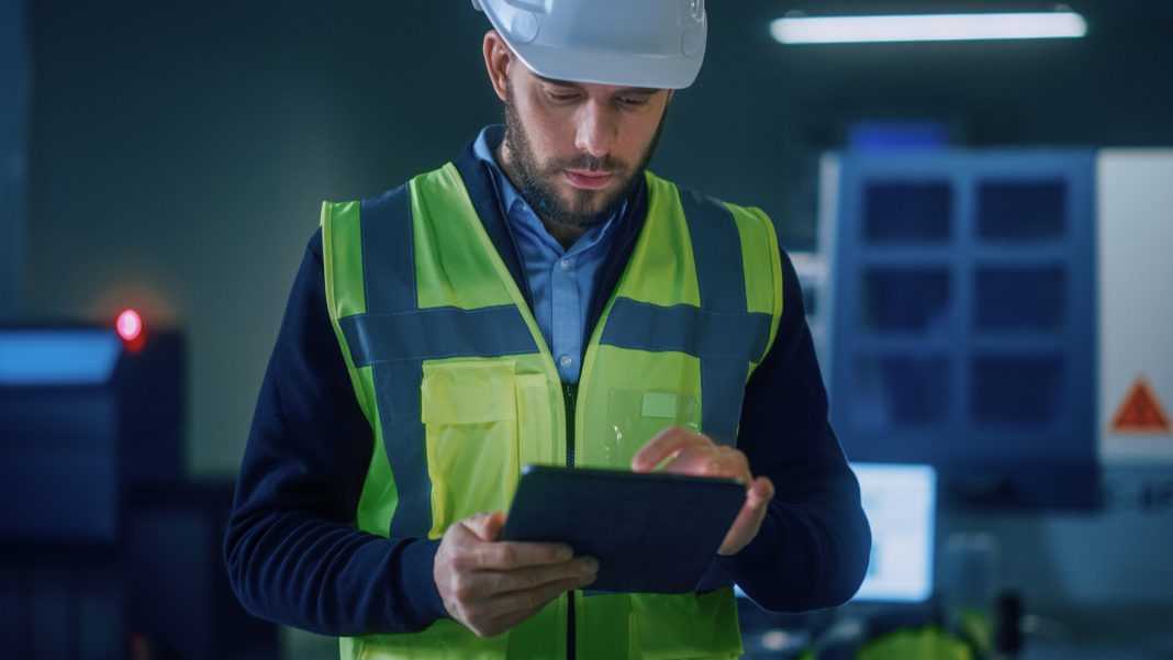 Professional Engineer Wearing Safety Vest and Hardhat Stands in Modern Factory Workshop, Uses Digital Tablet Computer. Handsome Professional Working in Industrial Facility with High Tech Machinery