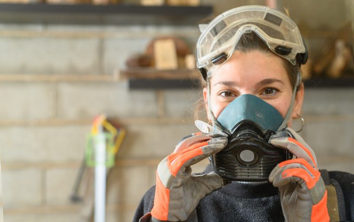 Woman in protective transparent googles and respirator looking at camera while standing in light workshop with professional equipment during woodwork, representing the need for respiratory protective equipment fit testing
