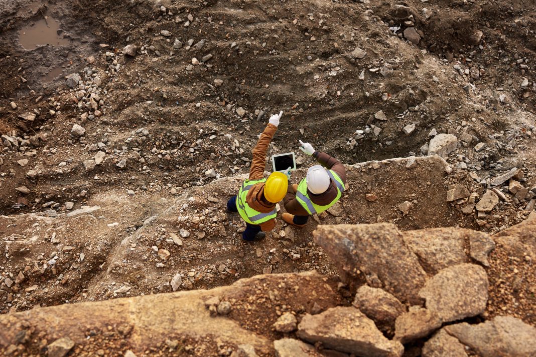 Top view shot of two industrial workers wearing reflective jackets standing on mining worksite outdoors using digital tablet, copy space, representing total productive maintenance