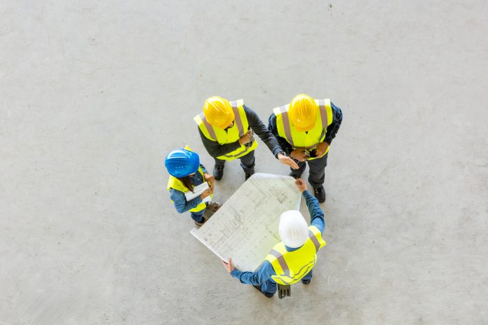 High angle view of architect, electrician and builders wearing work helmets and reflective vest standing on concrete floor, discussing over blueprint, representing the planning sector