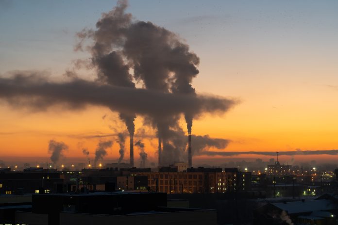 Chemical factory chimneys with raising smoke against red sunset sky in winter city during strong frost. View from afar of thermal power plant pipes emitting hazardous toxic pollutants into atmosphere
