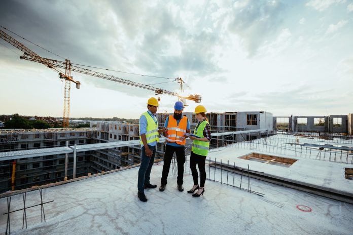 Employees working on construction site, wearing protective equipment and discussing next construction phase - full length models visible on the image. Representing head of responsible business