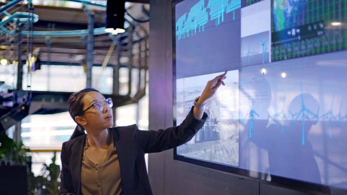 Stock photograph of a young Asian woman conducting a seminar / lecture with the aid of a large screen. The screen is displaying data & designs concerning low carbon electricity production with solar panels & wind turbines. These are juxtaposed with an image of conventional fossil fuel oil production, representing R&D tax relief