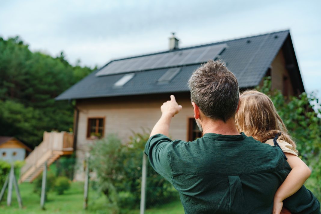 Rear view of dad holding her little girl in arms and showing at their house with solar panels, representing increased renewable energy installations in UK homes