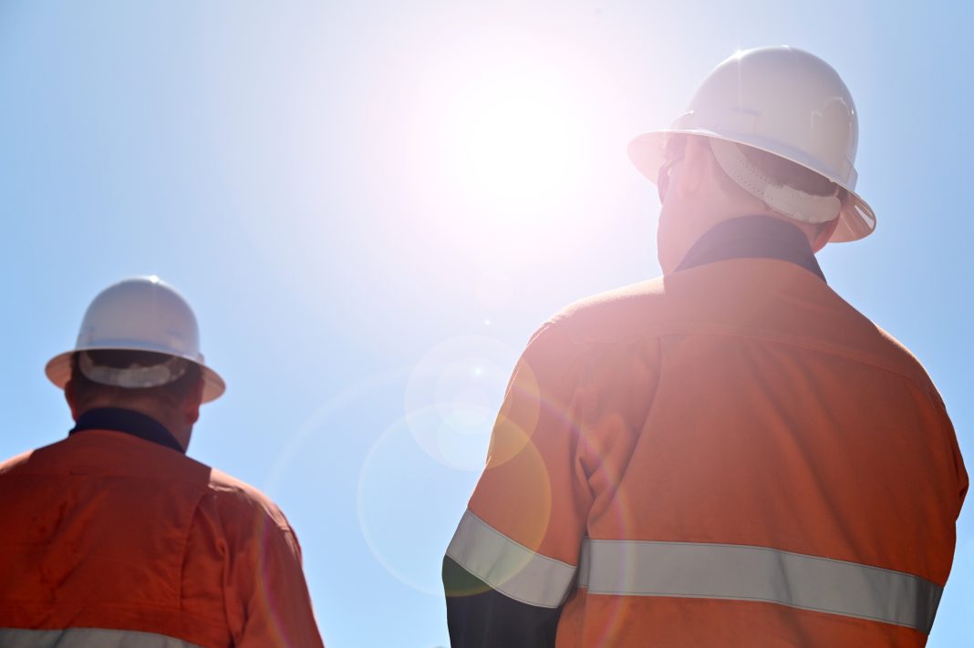Two unrecognizable blue collar workers isolated against clear blue sky with sun flare, representing the rise in monthly construction output