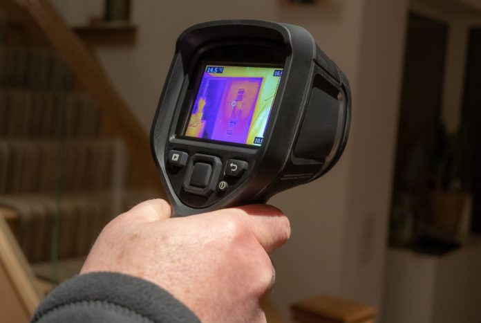 A male surveyor using a handheld infrared thermal camera, to inspect the interior of a domestic home, looking for heat anomalies. The camera viewfinder shows a coloured thermal image with varying temperature ranges, representing topics covered in the Actis- LABC CPD partnership