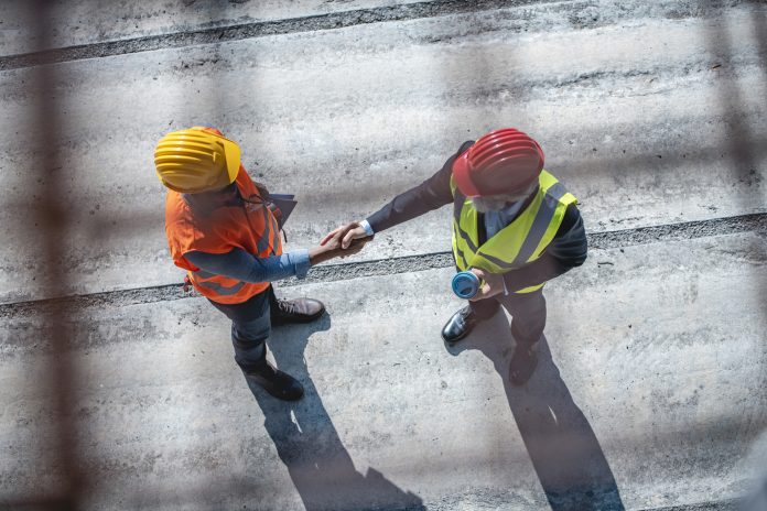 High angle view of young African construction site foreman and mature Caucasian project manager wearing reflective vests and hardhats and shaking hands, representing the CLC RAAC expert panel