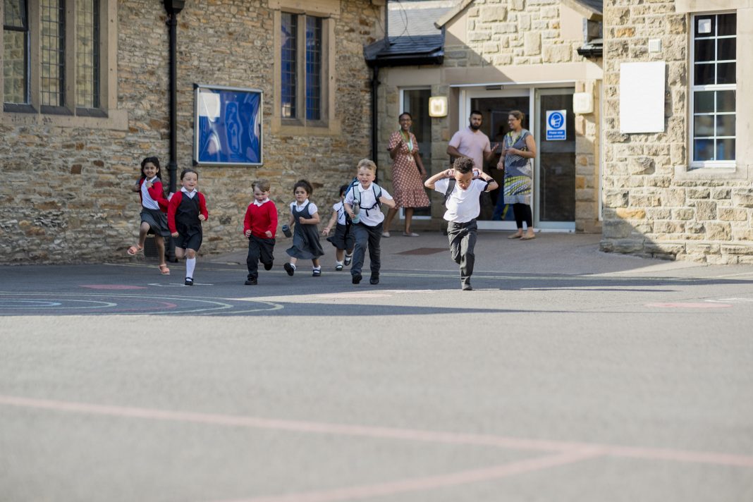 Children running in their school yard in the North East of England. They are all running away from the door with their backpacks on. Their teachers are standing at the door watching. Image represents the risk of RAAC concrete in schools
