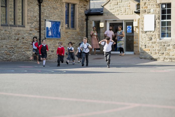 Over 100 schools told to evacuate due to RAAC concrete risk Children running in their school yard in the North East of England. They are all running away from the door with their backpacks on. Their teachers are standing at the door watching. Image represents the risk of RAAC concrete in schools