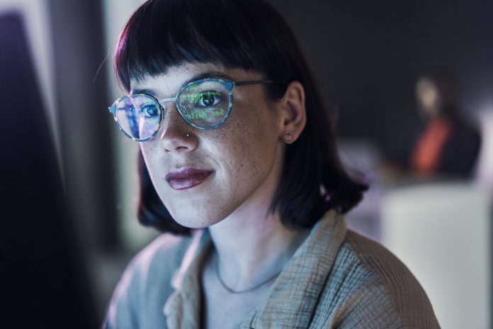 Business woman face, computer and ux coding of young employee with blue light and glasses. Digital code, hacker face and reading of a female worker at night planning with online cybersecurity data