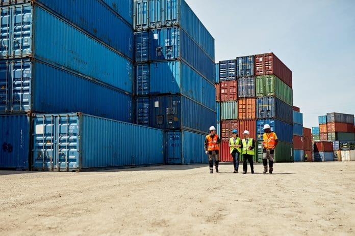Logistics Team Walking Together in Inland Port Mid distance view of diverse cargo handlers and supervisors approaching camera in reflective vests and hardhats amidst stacks of cargo containers.