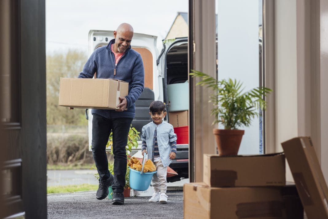 A shot of a grandfather with his grandson wearing casual clothing unloading boxes and furniture from the rear of a moving van, ready to move in to their new bungalow together. They are entering their new home through the door, representing Build Warranty's new home guide