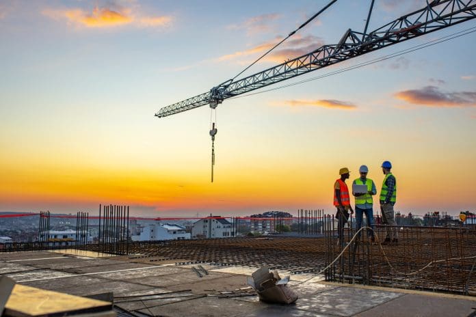 Photo of project manager visiting construction site with Multi-Ethnic construction workers standing on roof top at sunset. Business, building, industry, technology and people concept - smiling builder in hardhat with blueprint over group of builders at construction site under the tower crane, representing Watkin Jones
