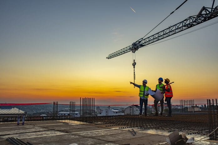Three Multi-Ethnic construction workers in uniform standing at construction site with crane in background, discussing building plans while holding blueprint at sunset under the tower crane.