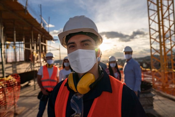 Portrait of a happy construction worker at a building site wearing a facemas with a group of workers at the background, representing mental health in construction