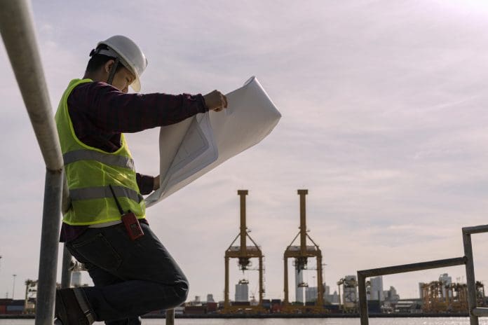 Engineer man wearing safety helmet and hold blueprints at a cargo port facility. Work outdoors Off-site exploration of the actual location representing construction materials in a circular economy