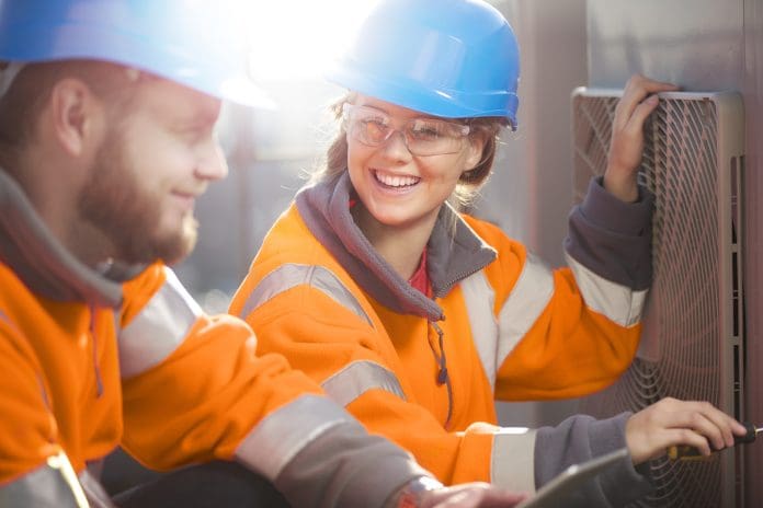 a female air conditioning technician with her supervisor on a roof top of an office block . They are performing routine servicing of the air conditioning units , and are wearing safety equipment , representing the Construction employability award