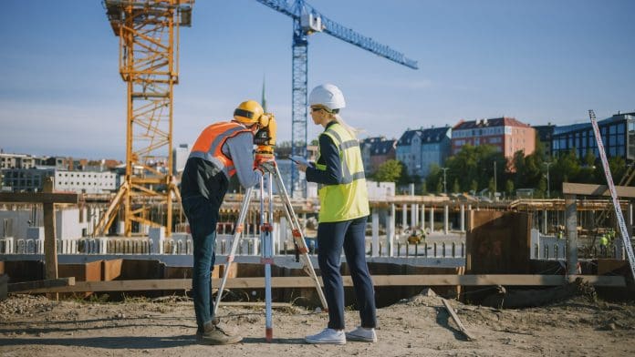Construction Worker Using Theodolite Surveying Optical Instrument for Measuring Angles in Horizontal and Vertical Planes on Construction Site. Engineer and Architect Using Tablet Next to Surveyor.