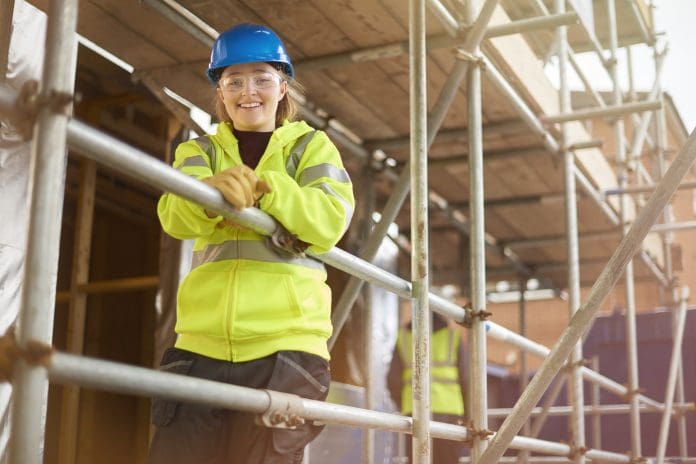 a female construction worker stands behind a scaffold and smiles to camera on a building site