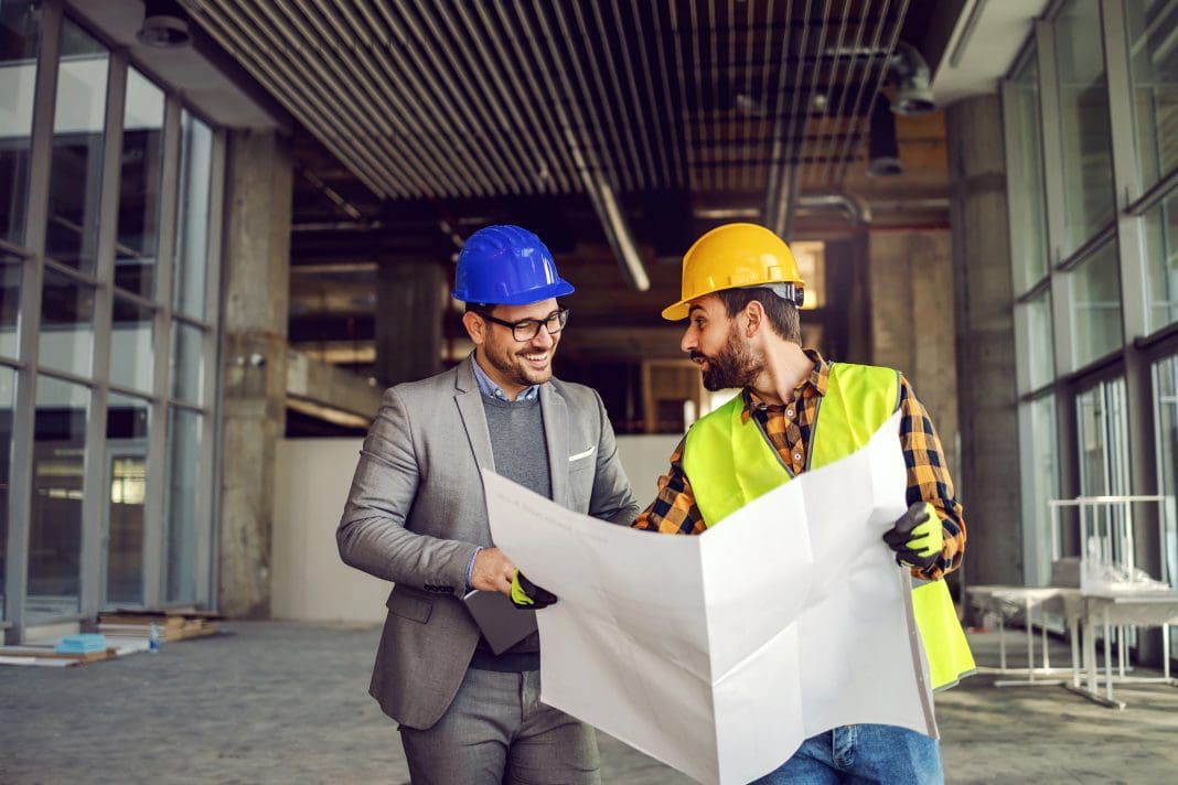 Smiling supervisor talking to construction worker about construction works. Worker holding blueprints and explaining to supervisor. Smiling supervisor talking to construction worker about construction works. Worker holding blueprints and explaining to supervisor, representing structural warranties