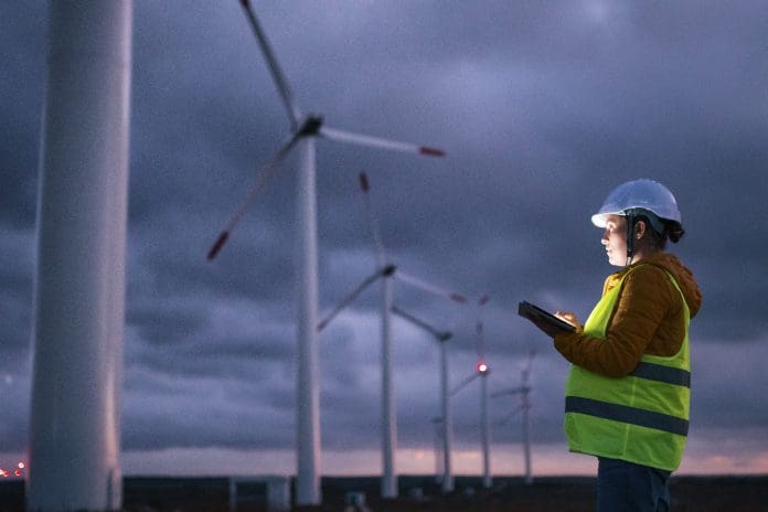 Electrical engineer working for the energy industry, supervising the condition of the Electrical Power Equipment in a wind turbines farm power station at night. Checking the data and the results of measurements with digital tablet. Pregnant woman engineer working on the field. Technology and Global Business.