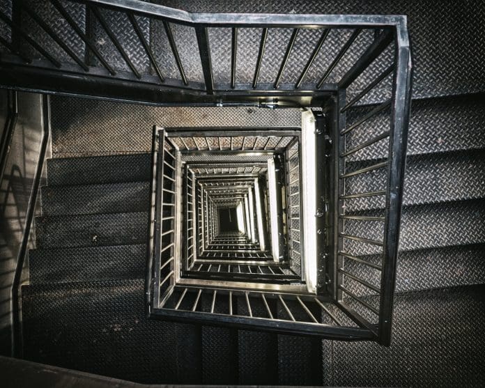 Government halts construction of 38,000 homes over second staircase delay A view from the top, looking down into an industrial building stairwell. Metal stairs and railings lead down several floors into the basement, representing the legal situation of a second staircase