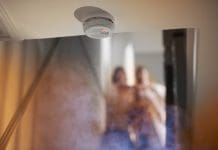 Landlords fined £20,000 for lack of fire safety and building regulation compliance smoke rises up towards a second floor in a two two storey house . Upstairs we can see a parent carrying a small child and running from a bedroom as they have been woken by the smoke alarm which is shown in focus at the edge of the ceiling leading to the stairwell.