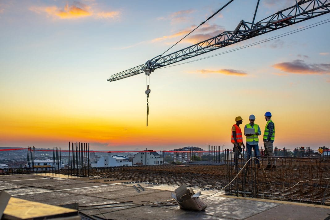 Photo of project manager visiting construction site with Multi-Ethnic construction workers standing on roof top at sunset. Business, building, industry, technology and people concept - smiling builder in hardhat with blueprint over group of builders at construction site under the tower crane.