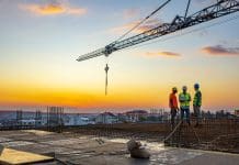 Newforma X PBC on-demand: Harnessing technology for AECO coordination Photo of project manager visiting construction site with Multi-Ethnic construction workers standing on roof top at sunset. Business, building, industry, technology and people concept - smiling builder in hardhat with blueprint over group of builders at construction site under the tower crane.