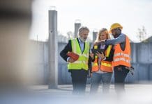 Newforma X PBC on-demand: Navigating AECO collaboration technology for construction Mature construction site managers meeting with young foreman and checking planning notes on digital tablet, collaboration technology