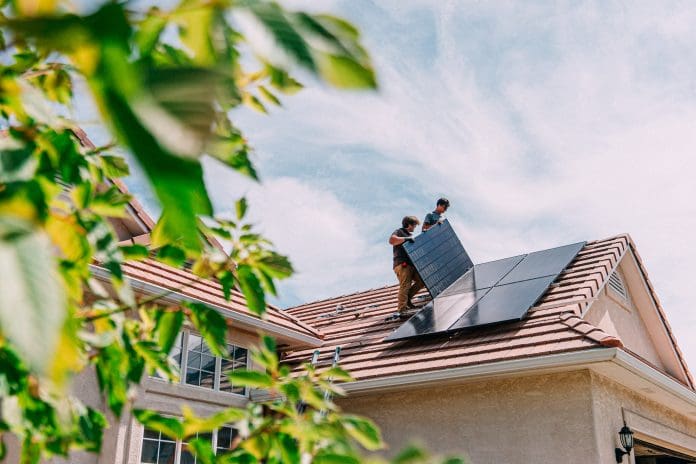 Low Angle view of Two Caucasian Male Homeowners or DIY-ers installing Solar Panels to Brackets on a Suburban Home with Clay Tiles in the Summer, representing the future homes standard