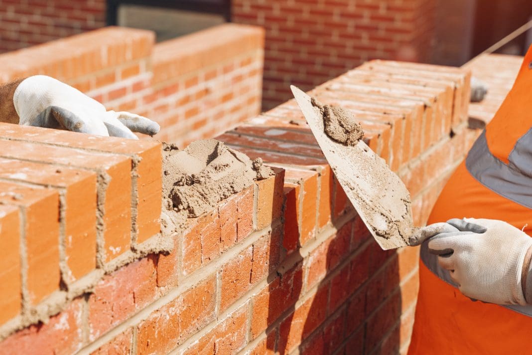 hand holding Masonry Trowel in protective gloves of bricklayer laying bricks at construction site The Brick Development Association (BDA) and Ceramics UK (C-UK) have made the decision to merge as a collective trade organisation.