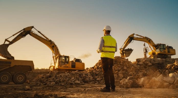 Caucasian Male Urban Planner Wearing Protective Goggles And Using Tablet On Construction Site On A Sunny Day. Man Inspecting Building Progress. Excavator Loading Materials Into Industrial Truck, representing construction administrations